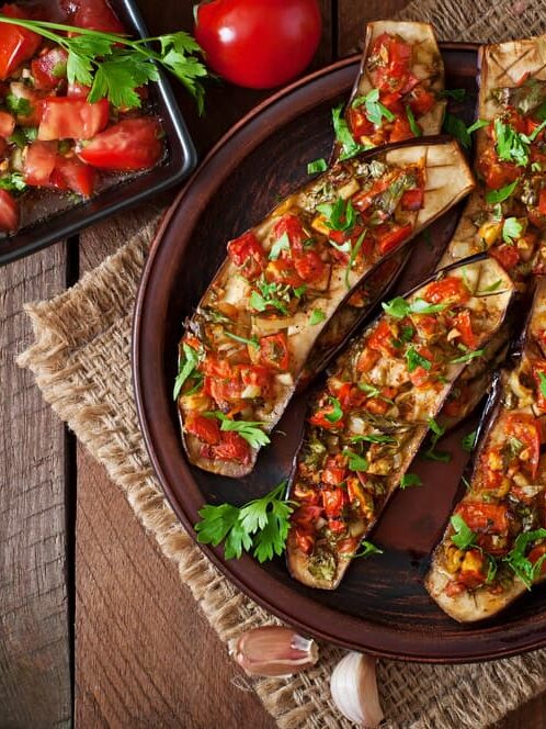 A vegan entre of toasted eggplant topped with semi dried tomatoes, onion and herbs stacked on a brown terracotta plate with garlic cloves on the hessian placemat beside it